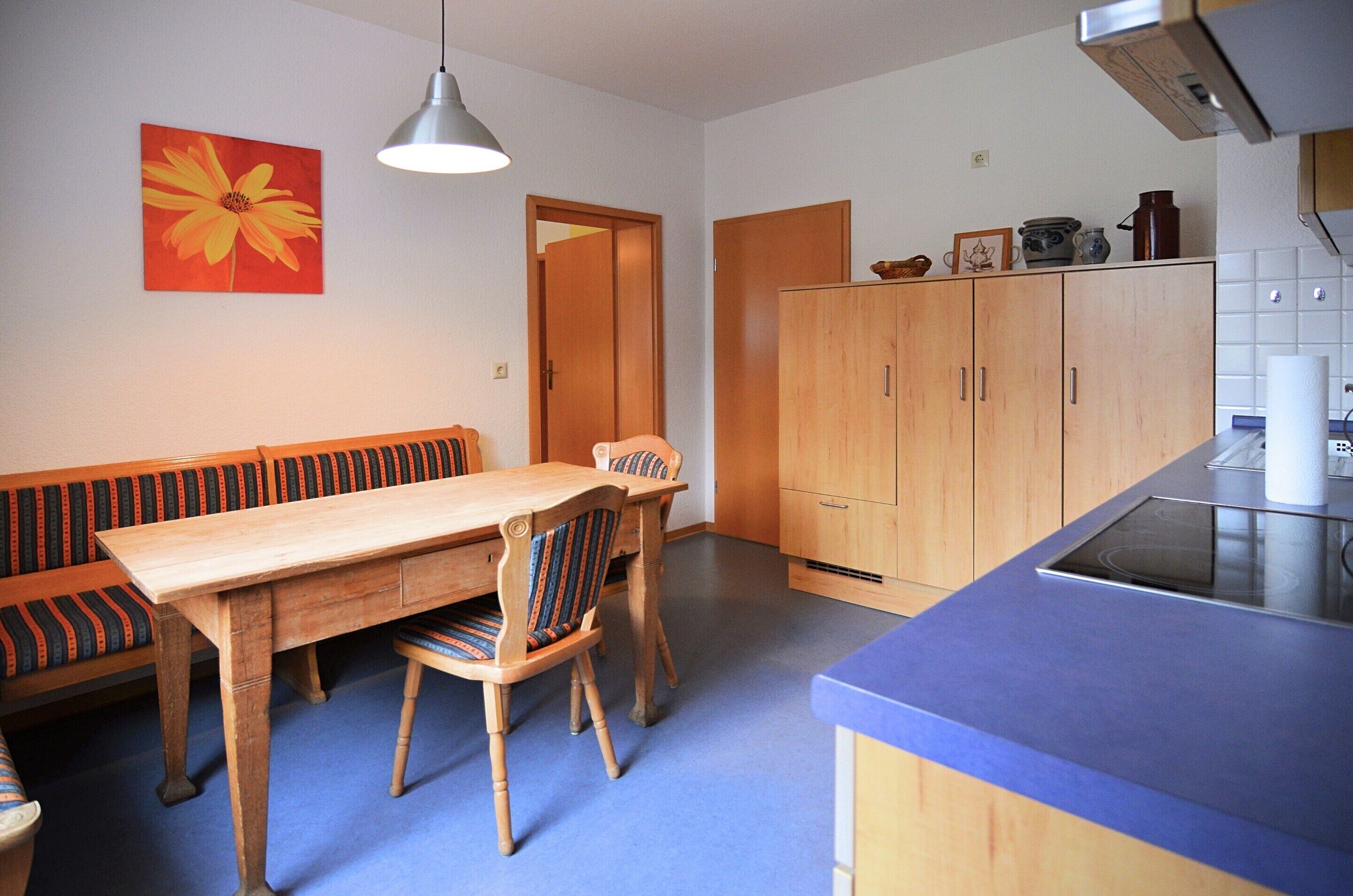 A dining area with a wooden table and chairs, a bench, and a kitchen with wooden cabinets and a blue countertop.