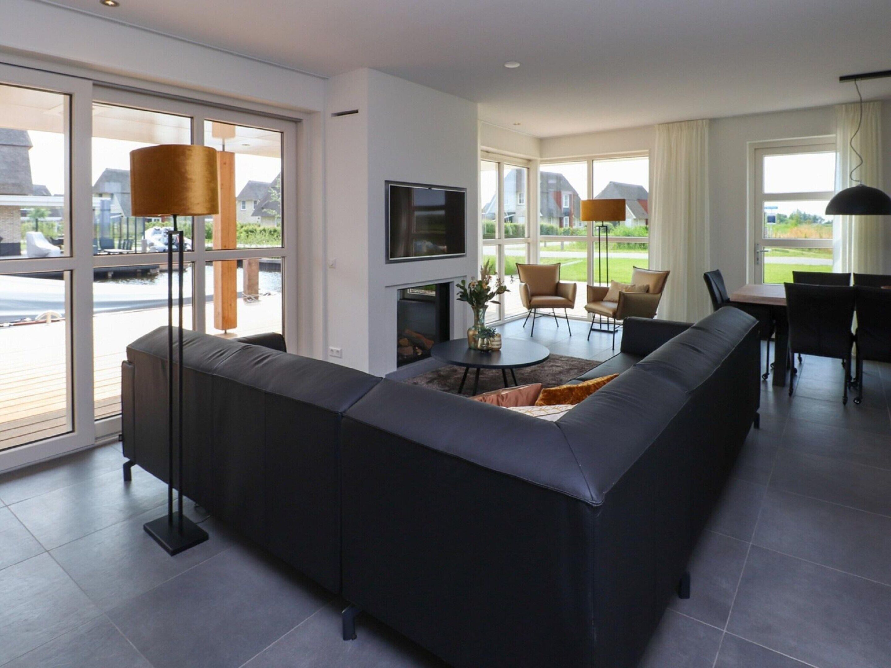 A modern living room with a large L-shaped sectional sofa, a round coffee table, and a flat-screen TV mounted on the wall. There are two floor lamps, a dining area with a table and chairs, and large windows offering a view of the outdoors.