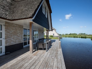Une maison avec un toit de chaume, une terrasse en bois et une vue sur une étendue d’eau paisible.