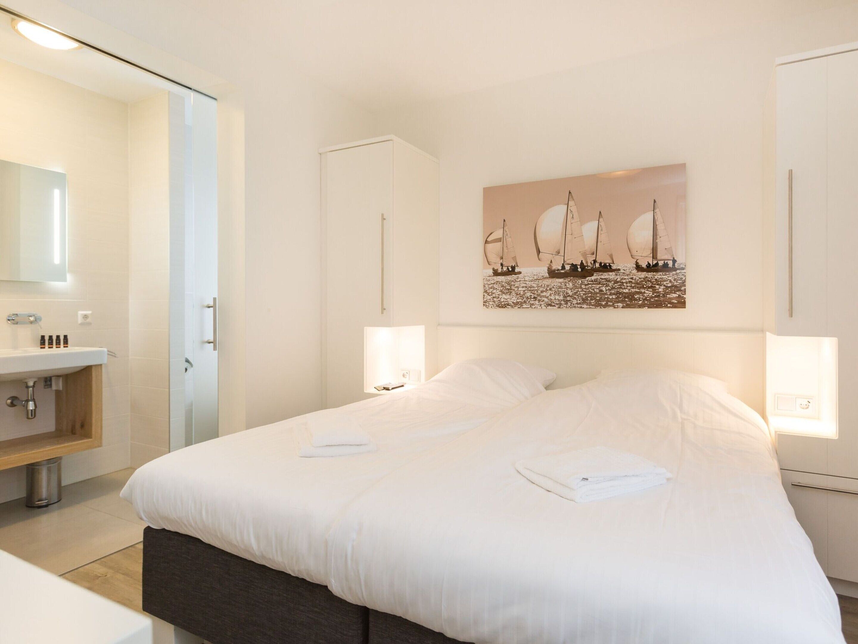 A hotel room with a large bed, a bedside table, a mirror, a sink, and a painting of sailboats on the wall.