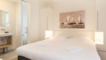 A hotel room with a large bed, a bedside table, a mirror, a sink, and a painting of sailboats on the wall.