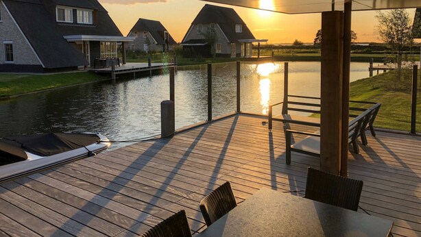 Une terrasse en bois avec des sièges extérieurs, donnant sur une étendue d’eau calme et sur des maisons en arrière-plan.