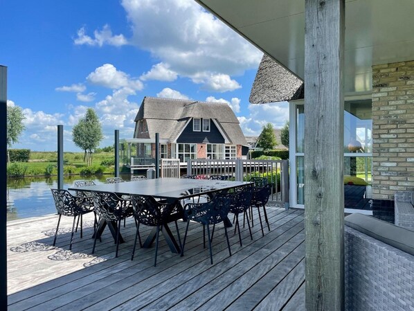 A wooden deck with a table and chairs, a thatched-roof building in the background, and a clear sky. - Luxury Villa With Sauna and Jacuzzi at the Lake (Delfstrahuizen)