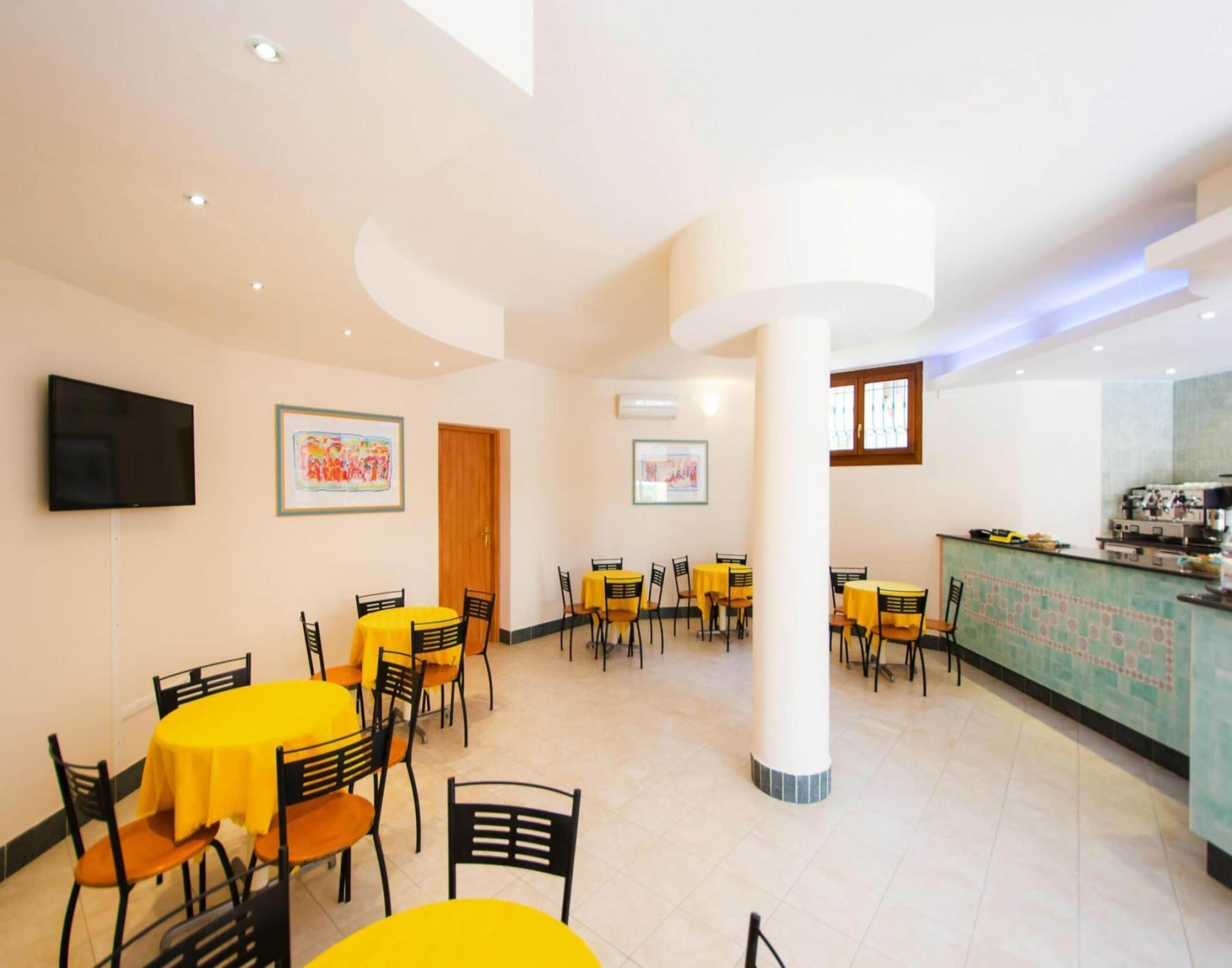 A dining area with yellow tables and black chairs, a television mounted on the wall, and framed artwork on the walls.