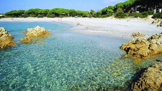Una spiaggia con acque limpide, formazioni rocciose e una riva sabbiosa sullo sfondo con vegetazione.