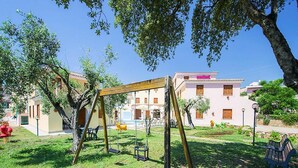 Un’area giochi con altalene e scivolo, circondata da erba e alberi, con un edificio sullo sfondo.