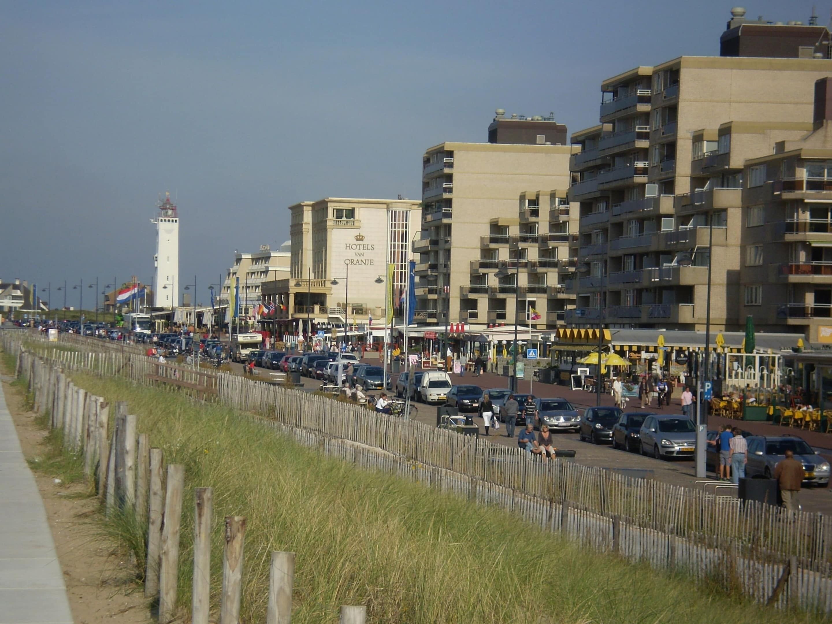 Un hôtel situé sur la côte, avec plusieurs étages, un phare imposant en arrière-plan et une rue animée en devant.
