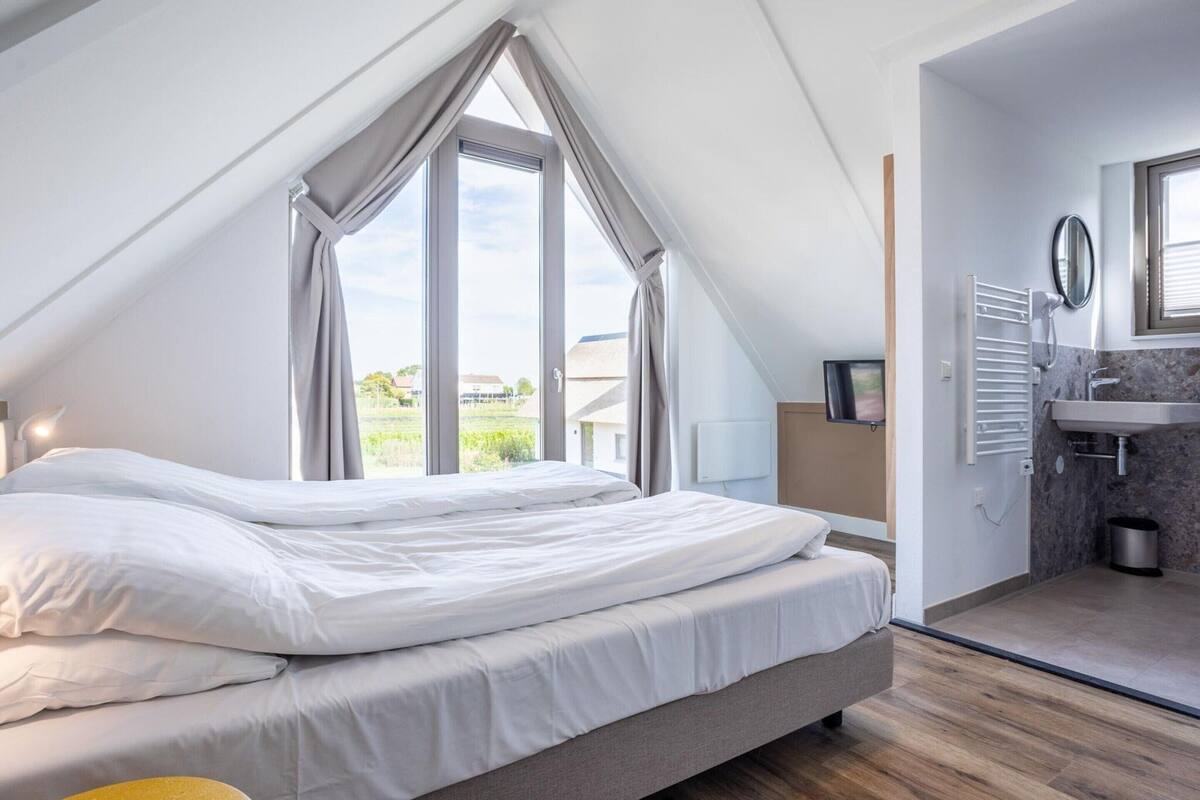 A bedroom with a large window, a bed, a TV, and a bathroom with a sink and mirror.
