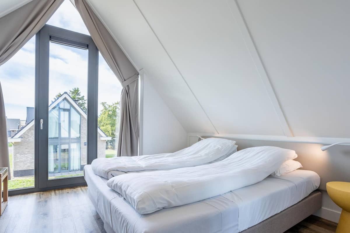 A bedroom with a large window, a bed, and a yellow stool.
