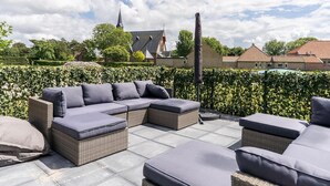 An outdoor seating area with wicker furniture and cushions, a hedge, and a view of a church and residential buildings in the background. - Lovely Apartment on Ameland Near Nes (Nes)