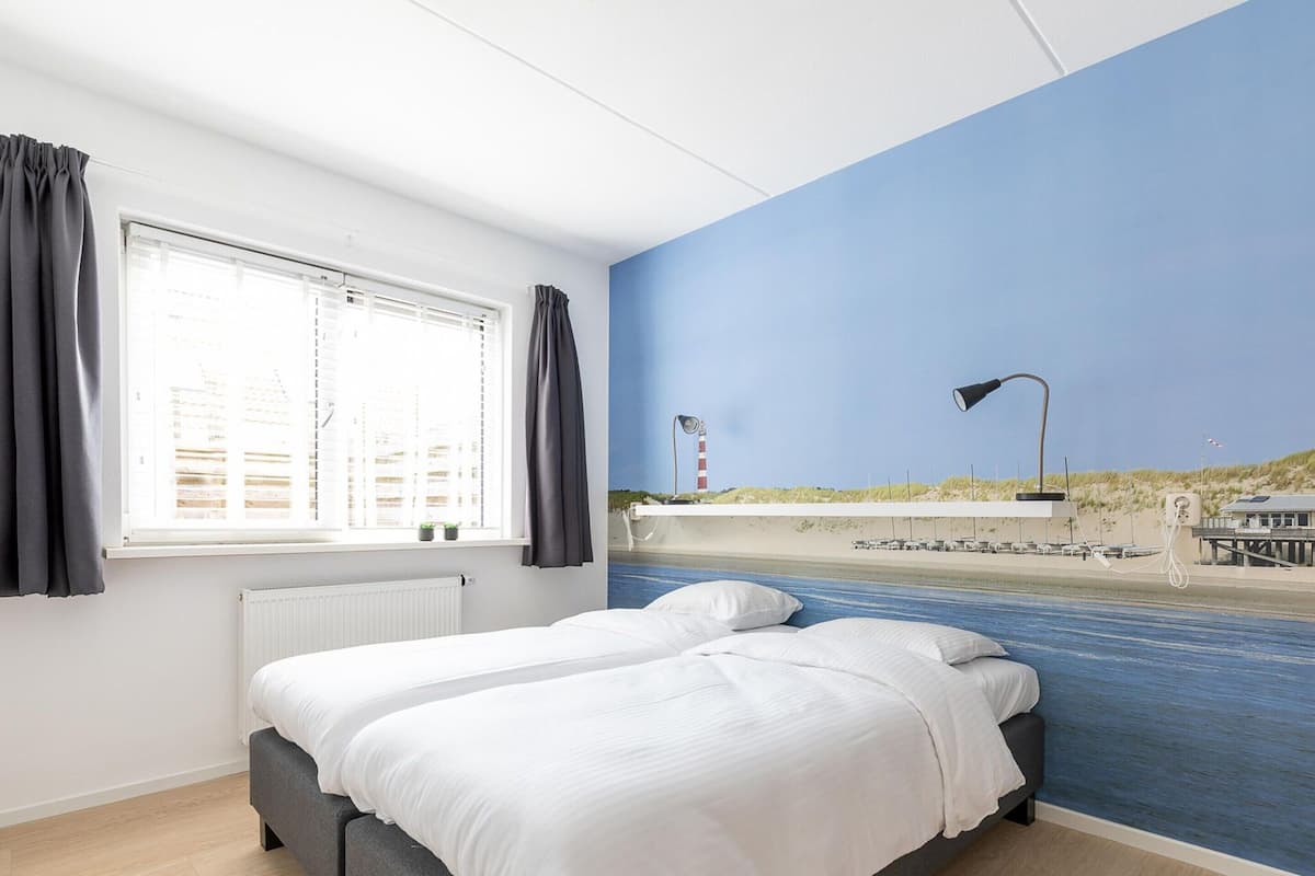 Ein Hotelzimmer mit einem Bett, einem Vorhangfenster, einem Heizkörper und einem Wandbild mit einer Strandansicht.