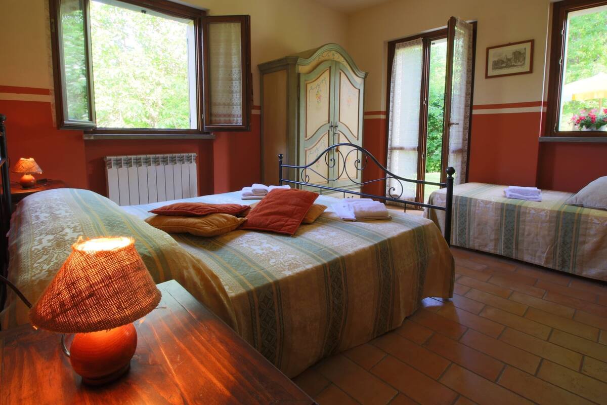 A bedroom with two beds, a wooden nightstand with a lamp, a radiator, and a window with curtains.
