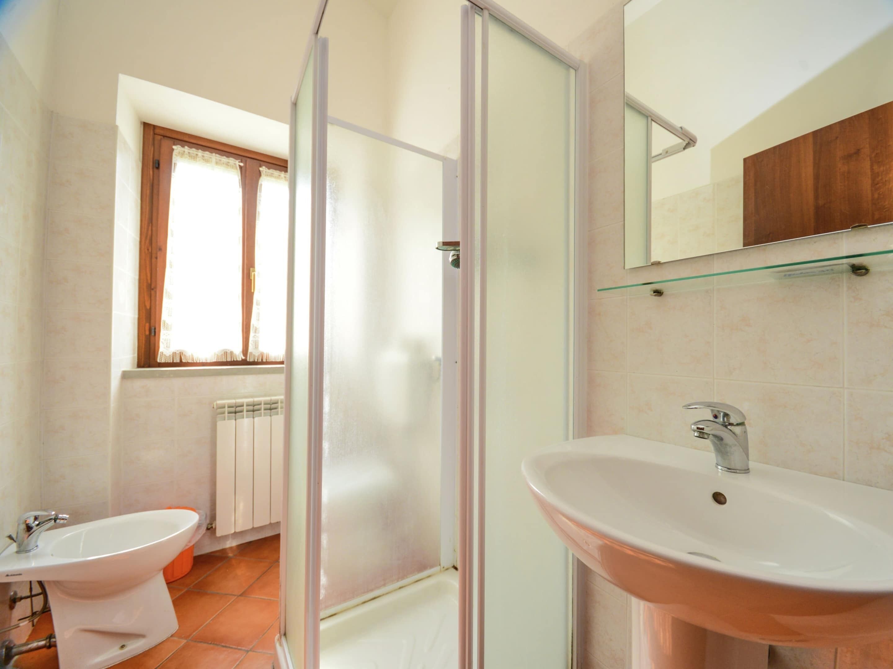 A bathroom with a glass shower enclosure, a sink, and a window with curtains.