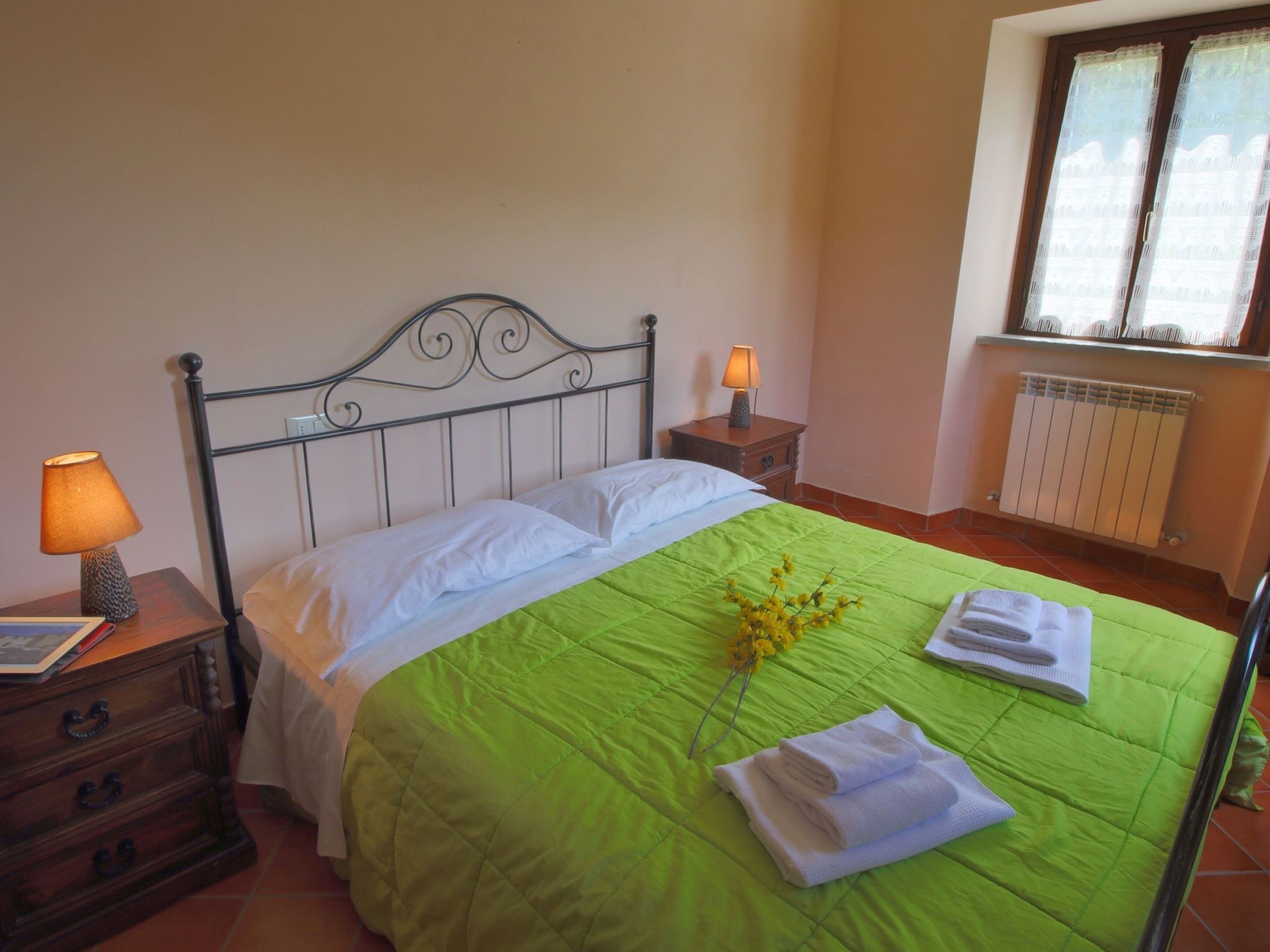 Un lit bien fait, recouvert d’une couverture verte, avec deux oreillers et un vase rempli de fleurs jaunes. Deux tables de chevet, chacune avec une lampe, sont placées de chaque côté du lit.