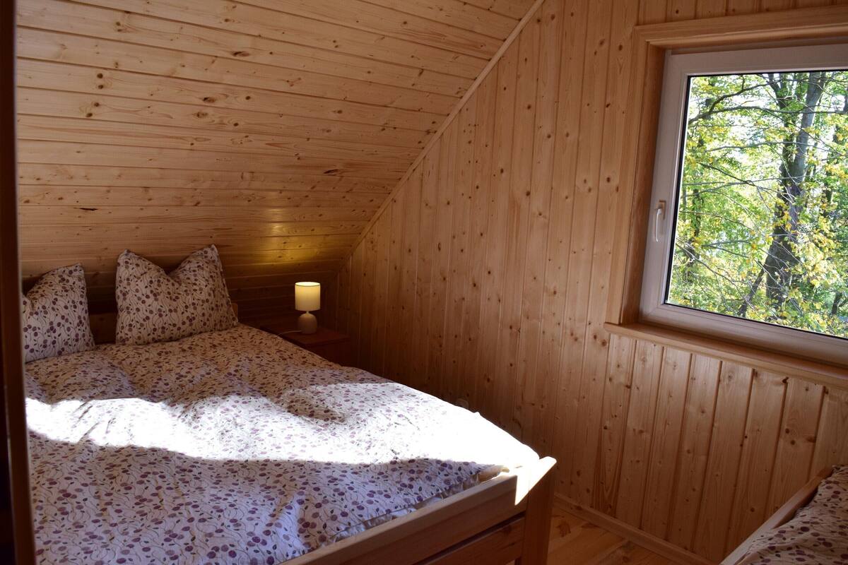 Ein Schlafzimmer einer Holzhütte mit Bett, Kissen, einem Nachttisch und einem Fenster mit Blick auf Bäume.