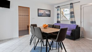 Un comedor con una mesa de madera, seis sillas y un bol de naranjas. Hay un sofá púrpura, una ventana con cortinas y un televisor de pantalla plana montado en la pared.
