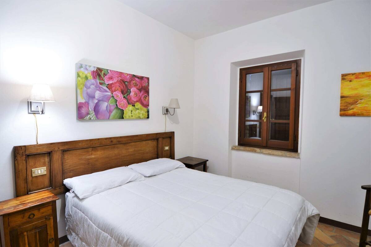 Une chambre à coucher avec une tête de lit en bois, un lit avec une literie blanche, une table de chevet avec une lampe, une fenêtre avec des cadres en bois et deux lampes fixées au mur.
