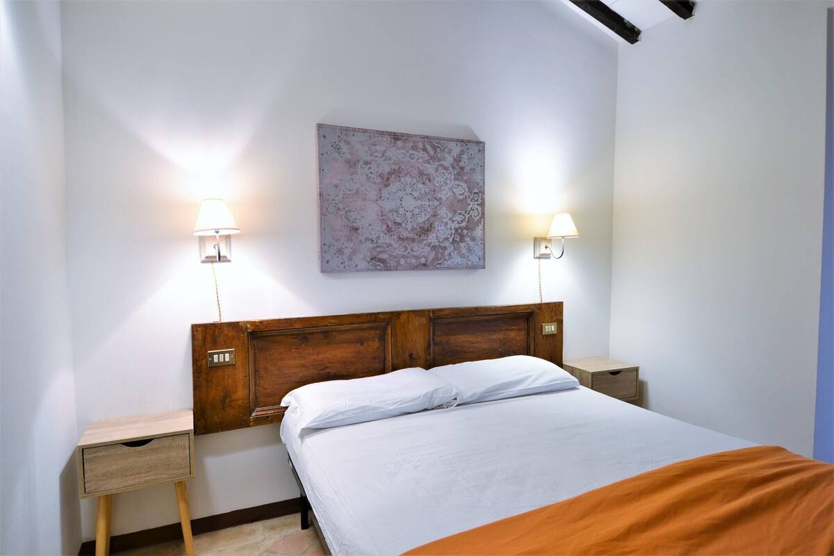Une chambre à coucher avec une tête de lit en bois, deux tables de chevet, une commode, un lit avec une literie blanche et deux lampes fixées au mur.