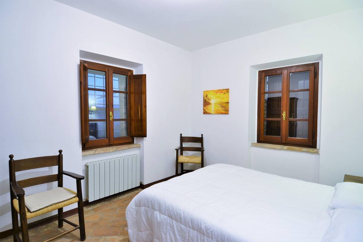 Une chambre avec deux fenêtres à cadre en bois, un lit avec une literie blanche, une chaise avec un coussin et un radiateur.