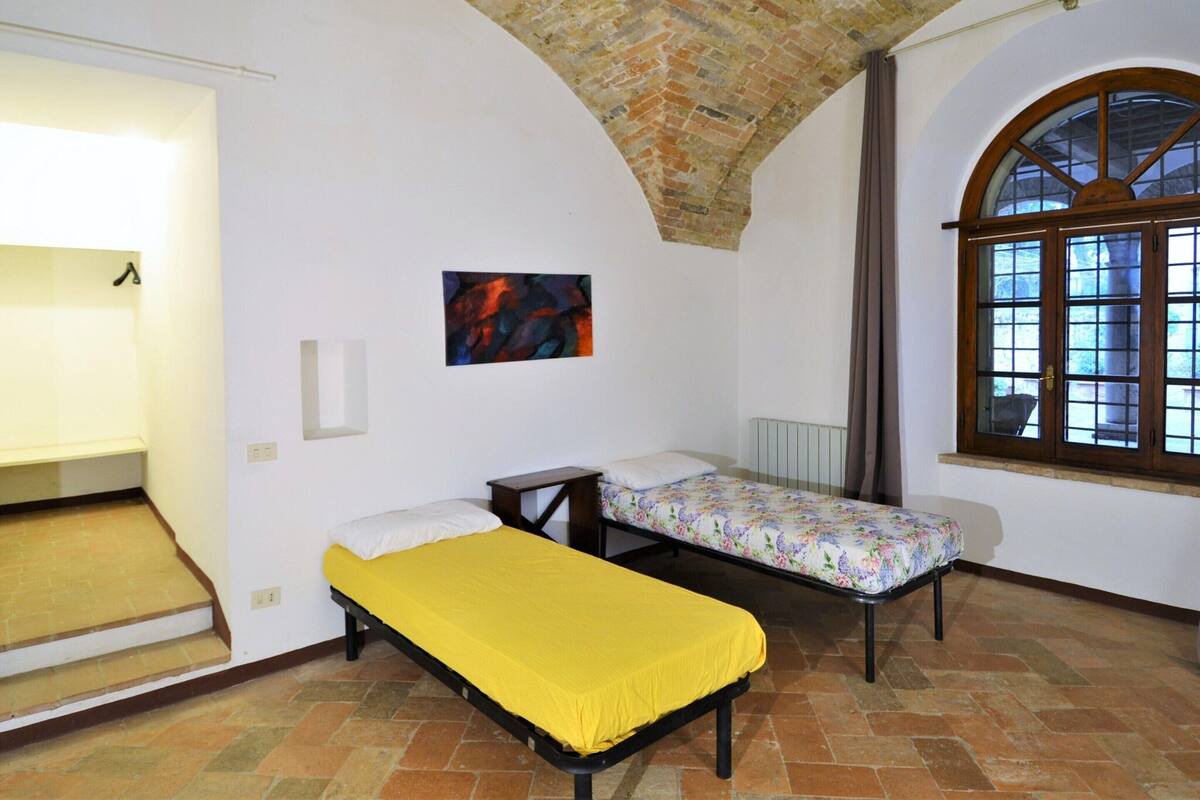Une chambre avec deux lits simples, une petite table, un tableau au mur et une grande fenêtre en arc de cercle.