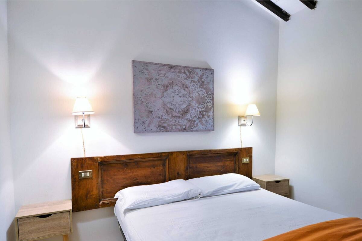 Une chambre à coucher avec une tête de lit en bois, deux tables de chevet, un lit recouvert de linge de maison blanc et un tableau abstrait accroché au mur.