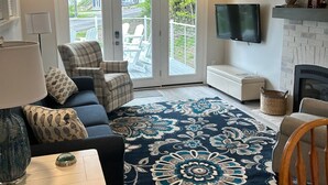 Een woonkamer met een bank, een fauteuil, een televisie, een open haard en uitzicht door glazen deuren.