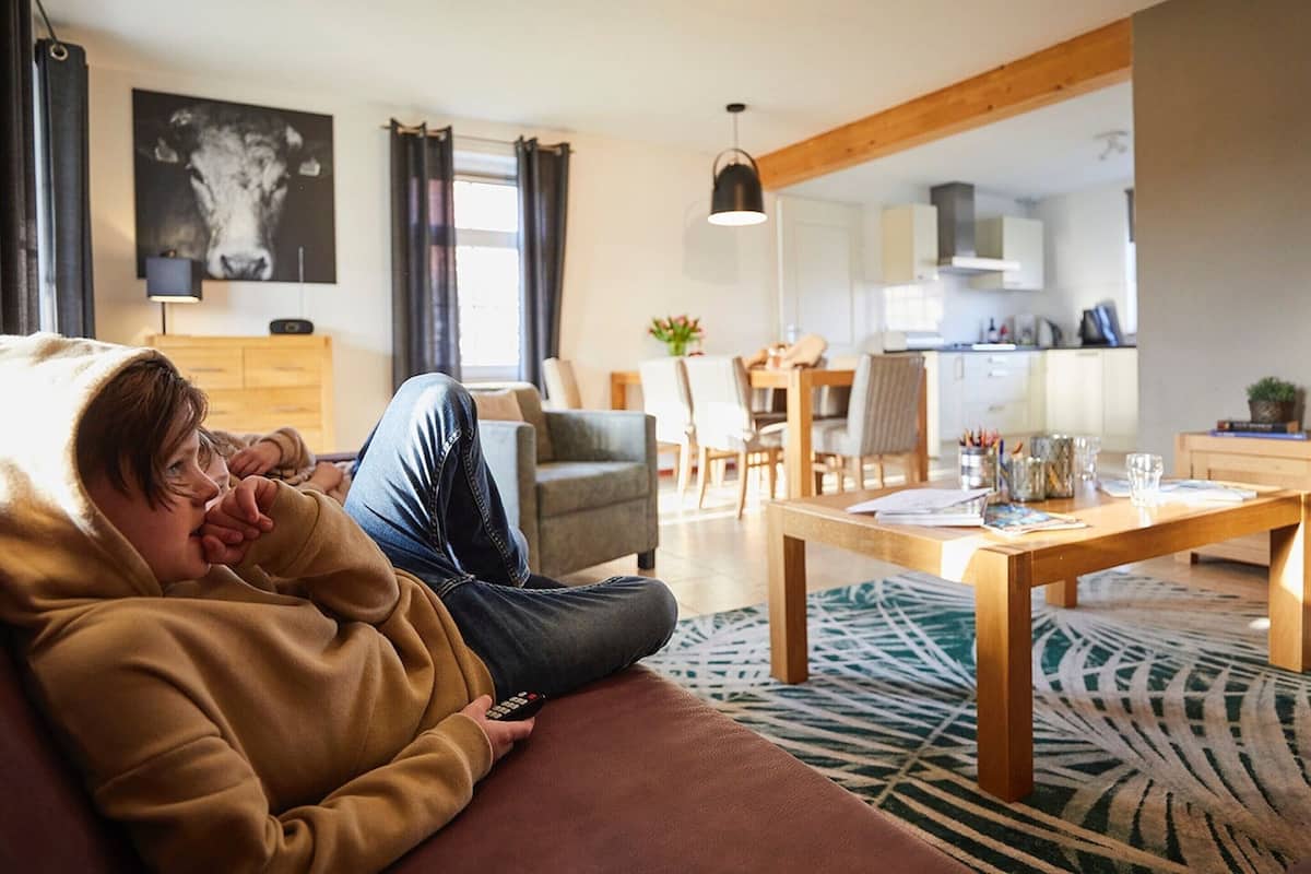 A person lounging on a sofa in a living room with a dining area and kitchen in the background.