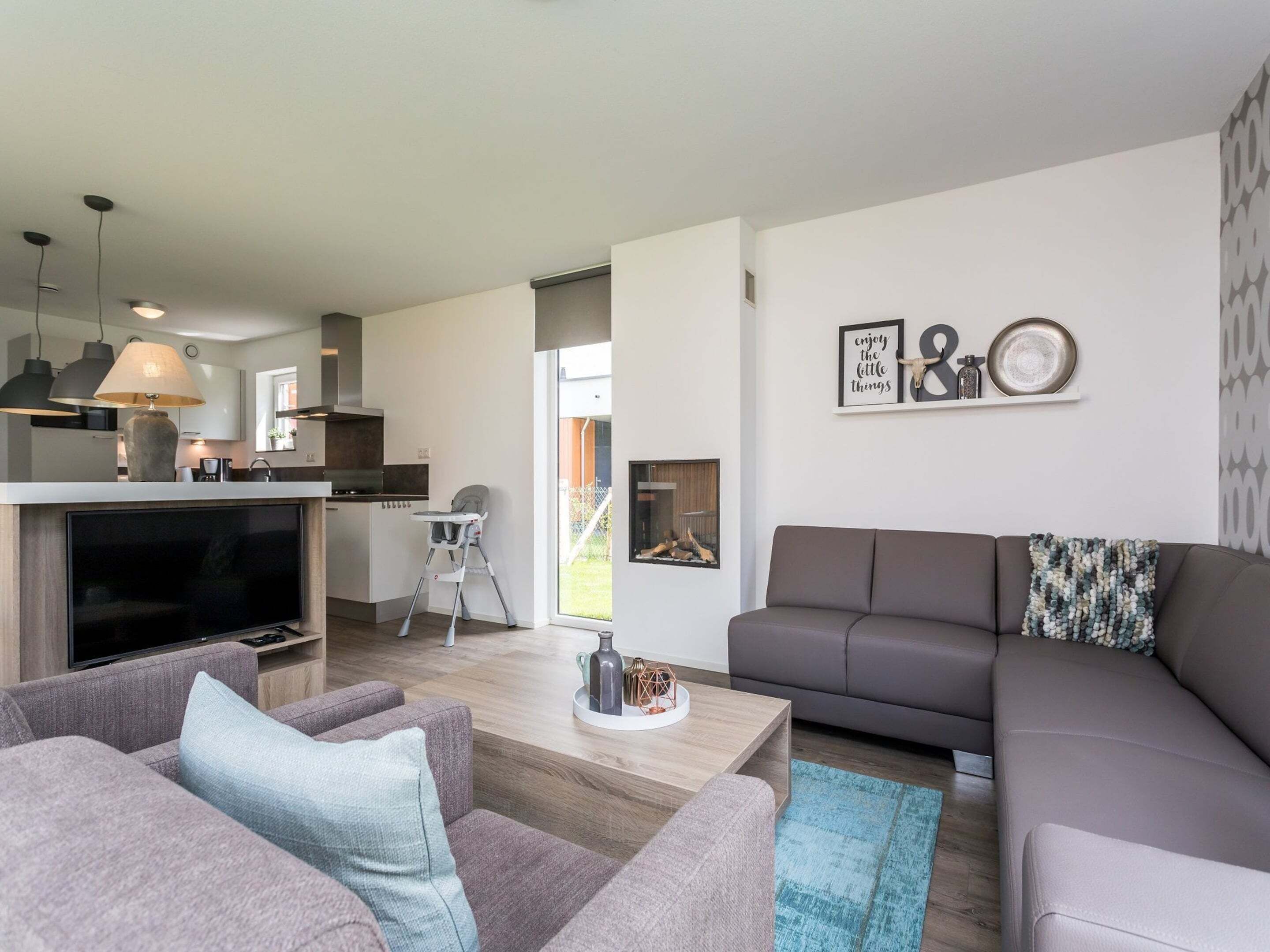 Una sala de estar moderna con un sofá gris, una mesa de centro y un televisor. Hay una zona de cocina con una mesa de comedor y sillas, además de una trona para bebé.