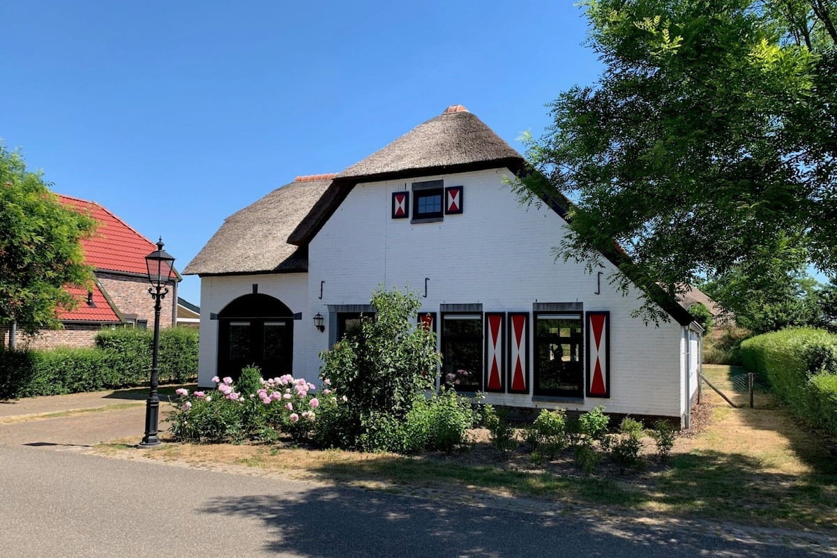 A white building with a thatched roof, black shutters, and a small garden with pink flowers.