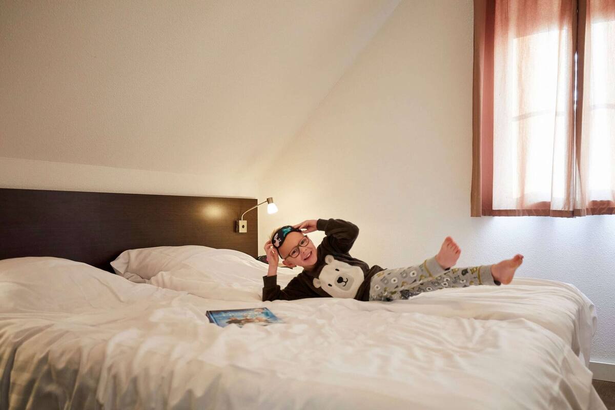 A child lying on a bed with a book, wearing pajamas with a bear design, in a room with a headboard and a window with curtains.