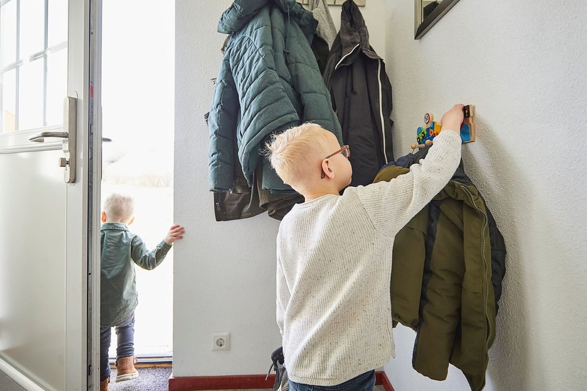 A child reaching for a toy on a wall-mounted shelf in a hallway with coats hanging on a rack.