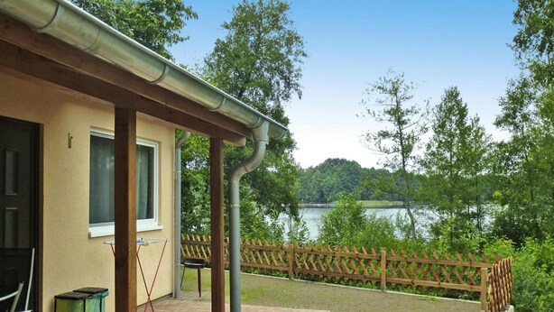 A patio with a brick floor, a wooden fence, and a view of a lake. - Feriendorf am Weinberg, Dobbrikow (Nuthe-Urstromtal)