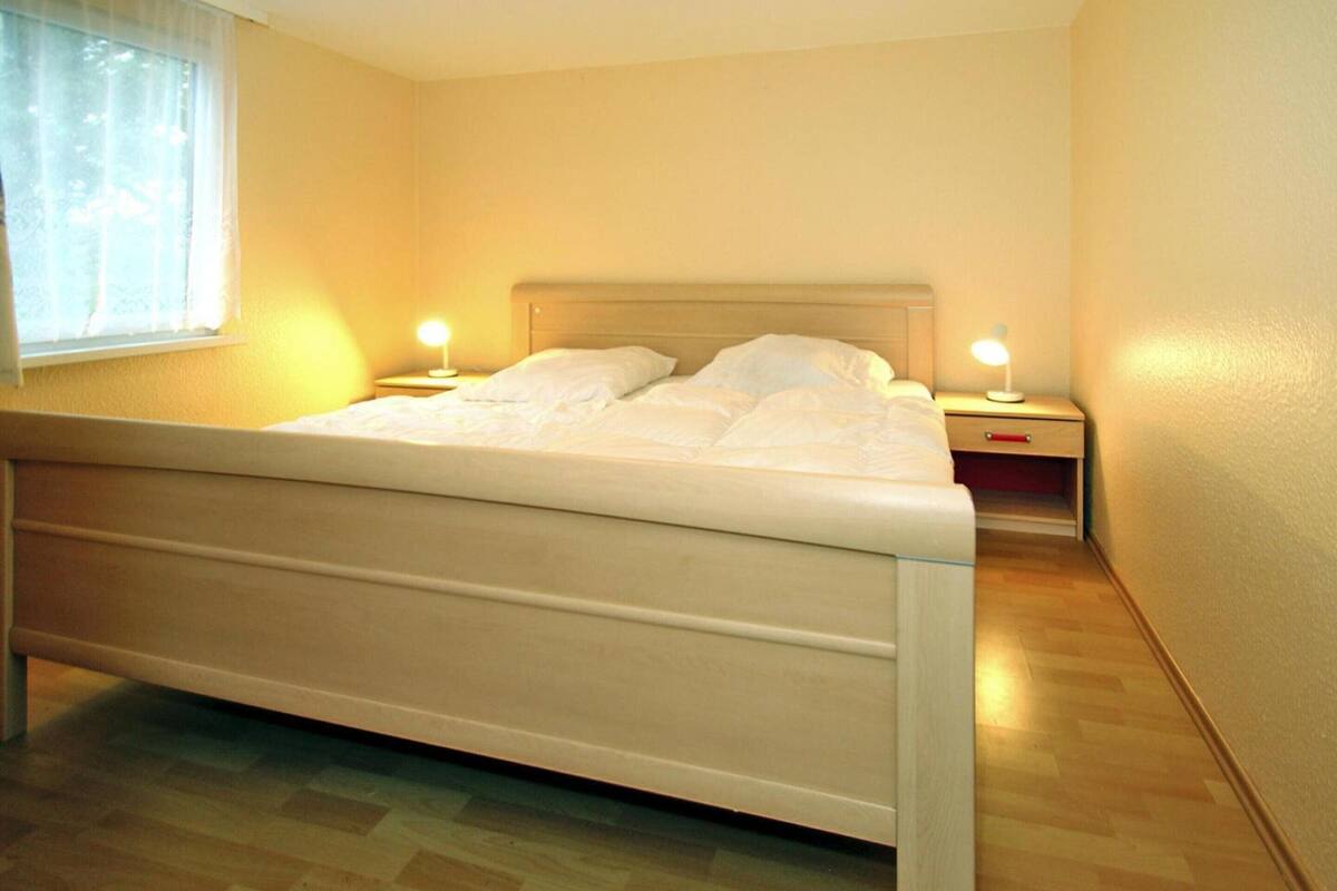 A neatly made bed in a room with wooden flooring and a window with sheer curtains.