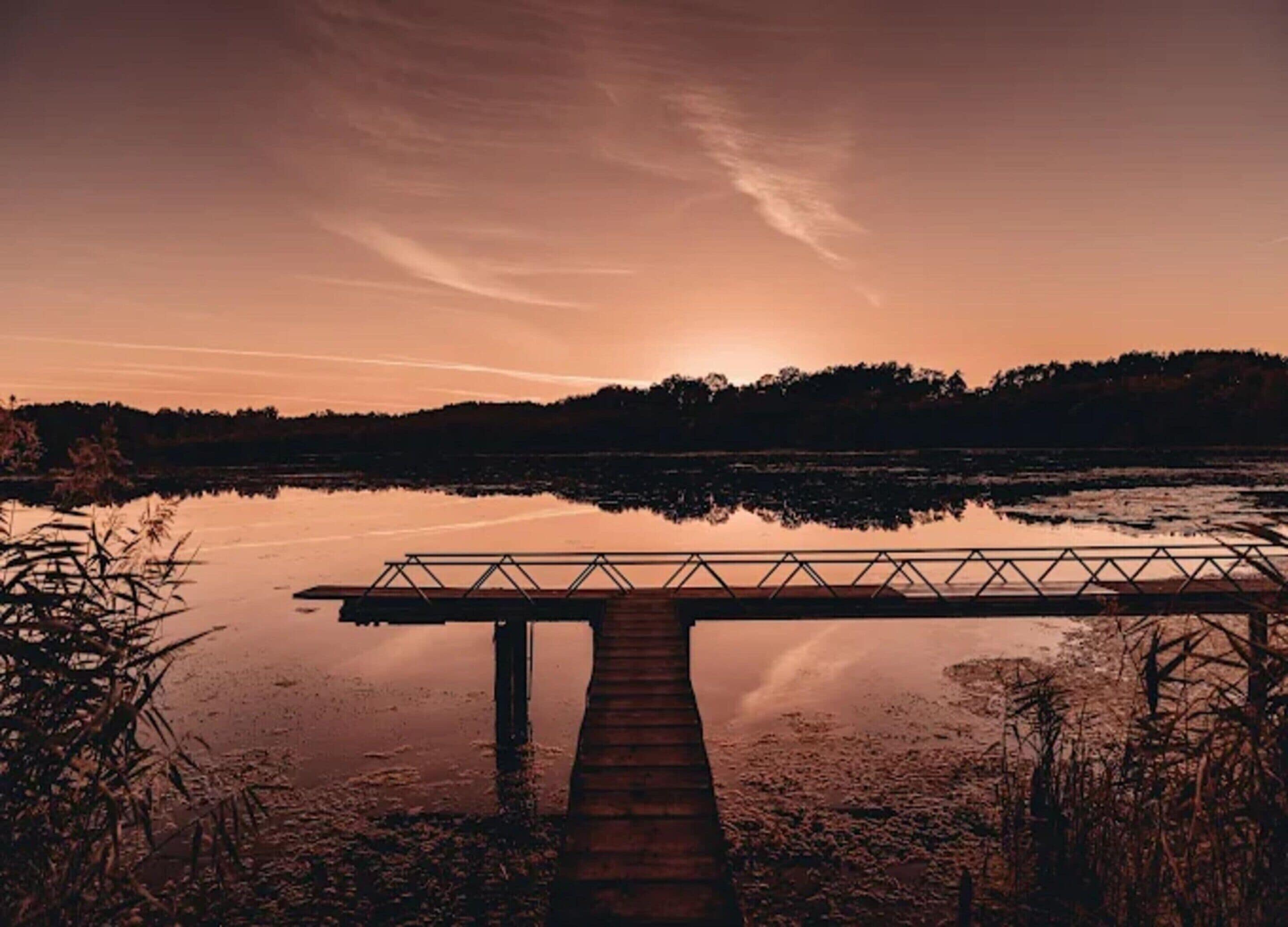 Un muelle de madera se adentra en una masa de agua tranquila, con un atardecer en el fondo.