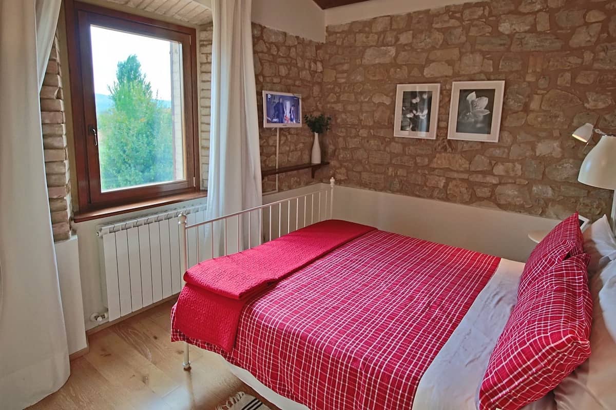 A bedroom with a stone wall, a window with curtains, a bed with a red and white checkered bedspread, and two framed pictures on the wall.