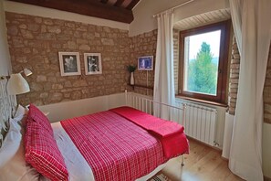 A bedroom with a stone wall, a bed with red bedding, a window with curtains, and framed pictures on the wall. - Modern Holiday Home in Foligno - Loc. With Pool (Foligno)