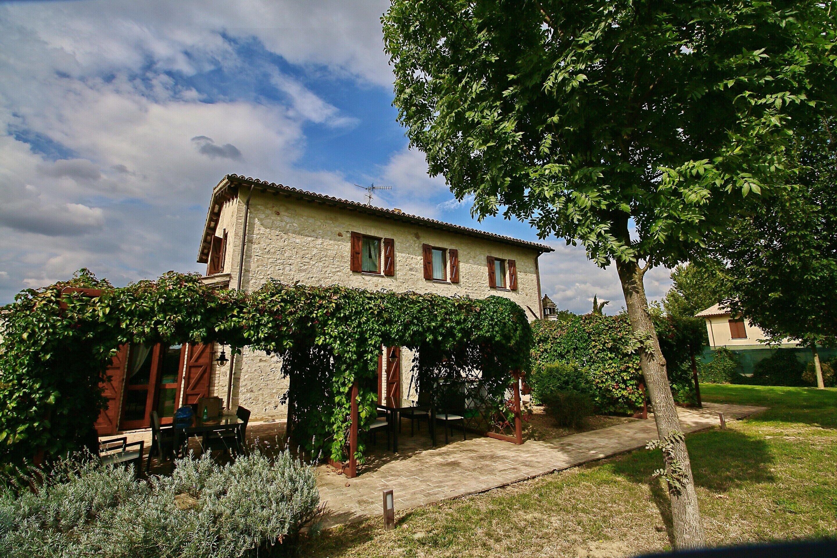 A stone building with a covered patio, a pathway leading to the entrance, and a garden area with shrubs.