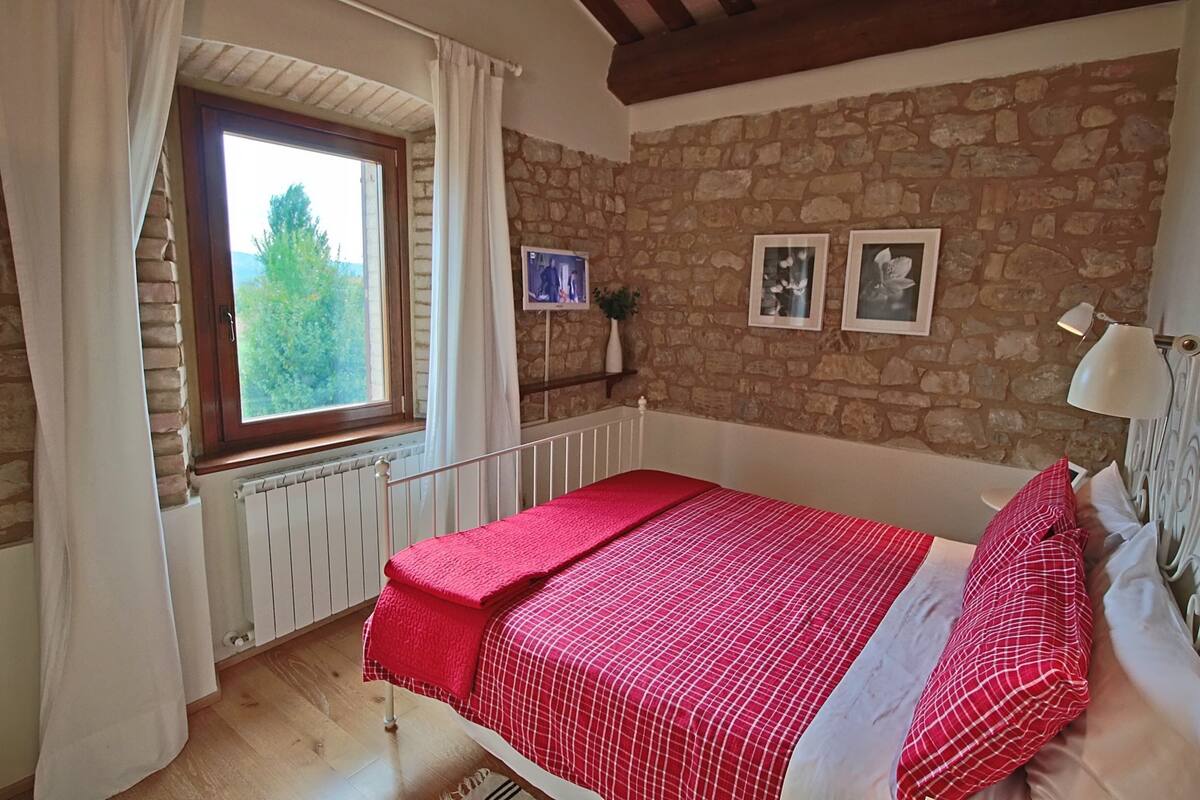 A bedroom with a stone wall, a window with curtains, a bed with a red and white checkered bedspread, and two framed pictures on the wall.
