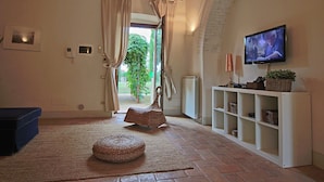 A room with a television, a wicker ottoman, a white shelving unit, and a view of greenery outside. - Modern Holiday Home in Foligno - Loc. With Pool (Foligno)