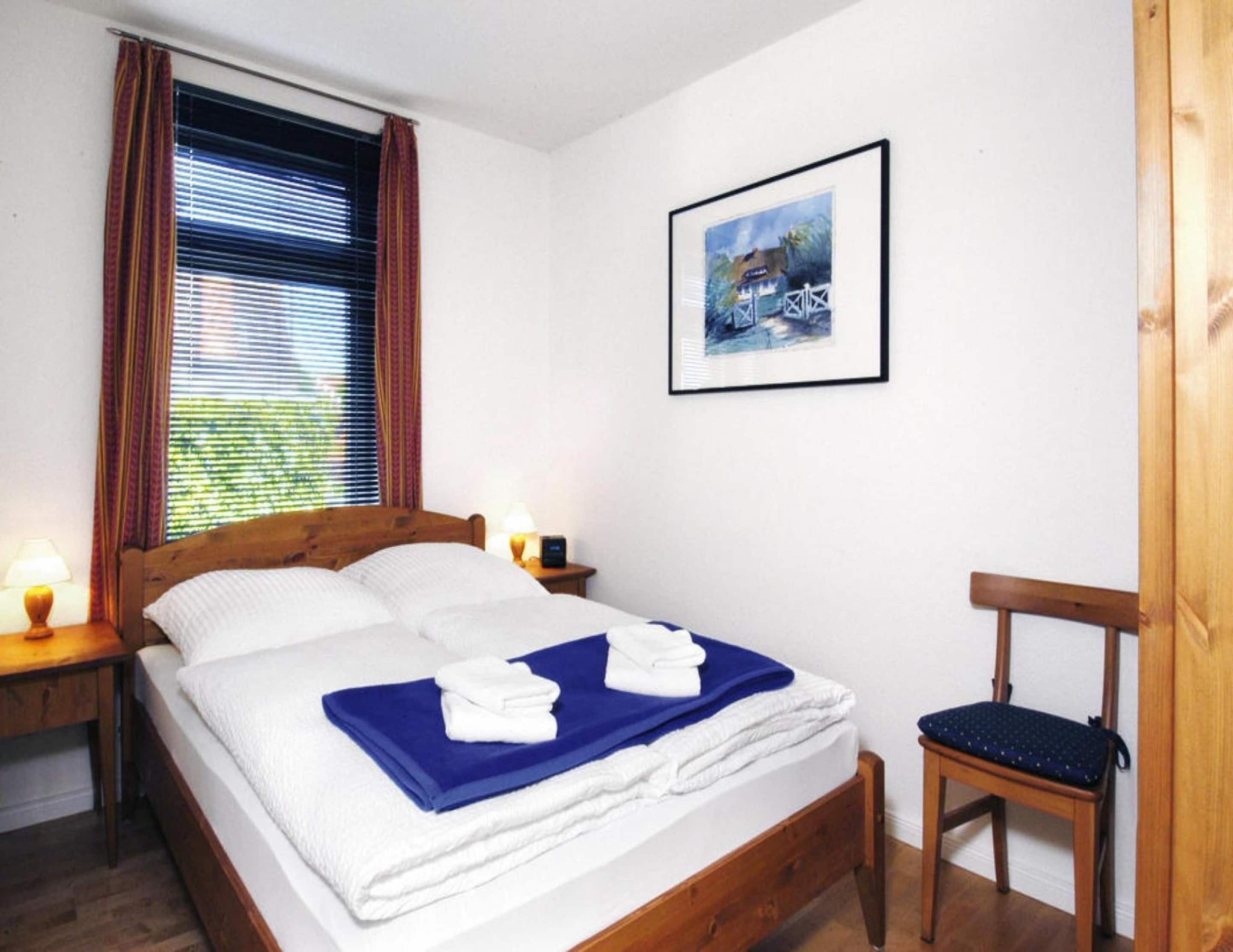 Een net opgemaakt bed met witte en blauwe lakens, een ingelijste foto aan de muur, en een houten stoel.