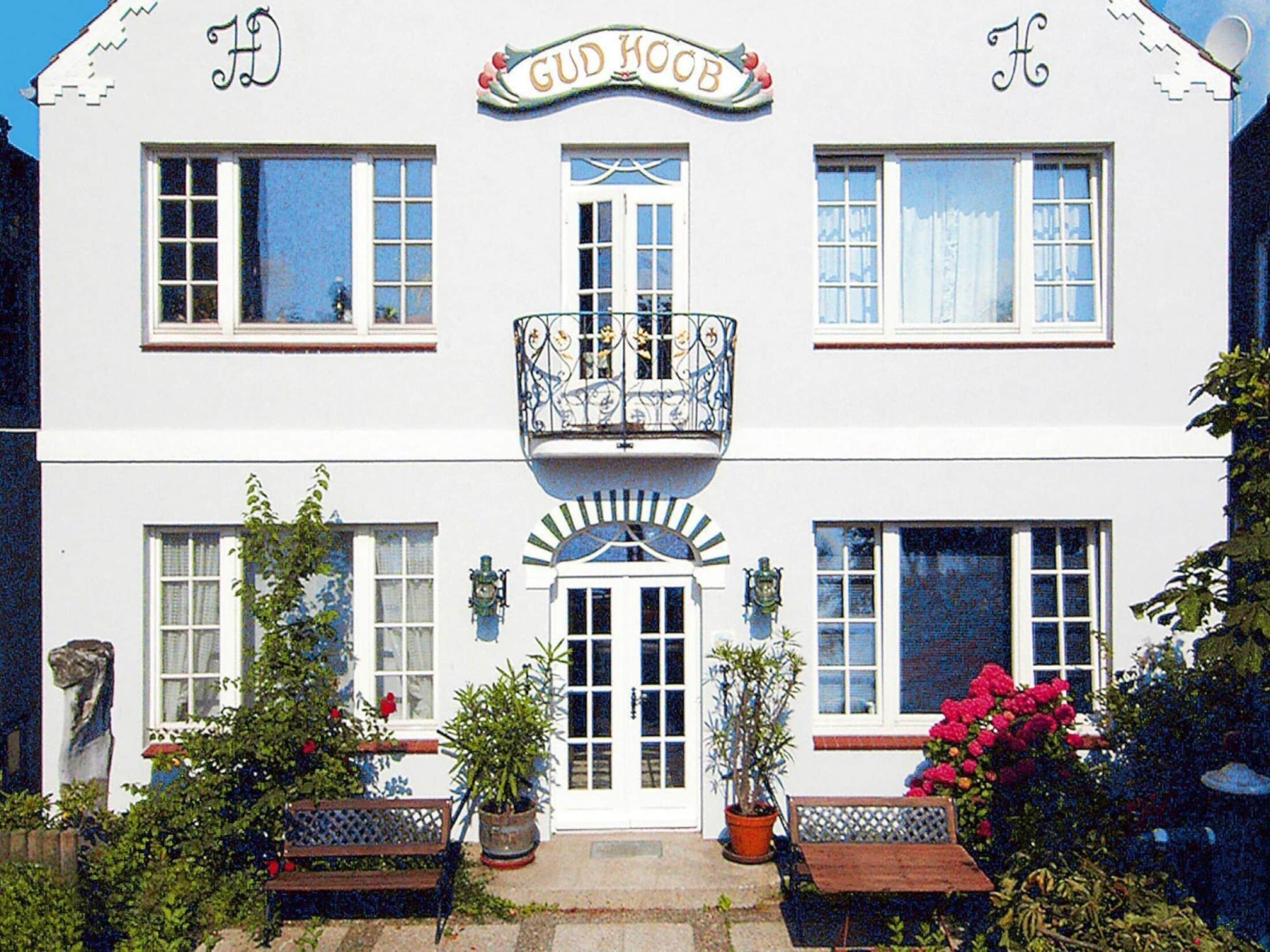 A two-story building with a sign reading "GUD HOOR". There are two windows on each floor, a balcony with ornate railings, and a small garden area with benches and potted plants.