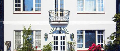 A two-story building with a sign reading "GUD HOOR". There are two windows on each floor, a balcony with ornate railings, and a small garden area with benches and potted plants.