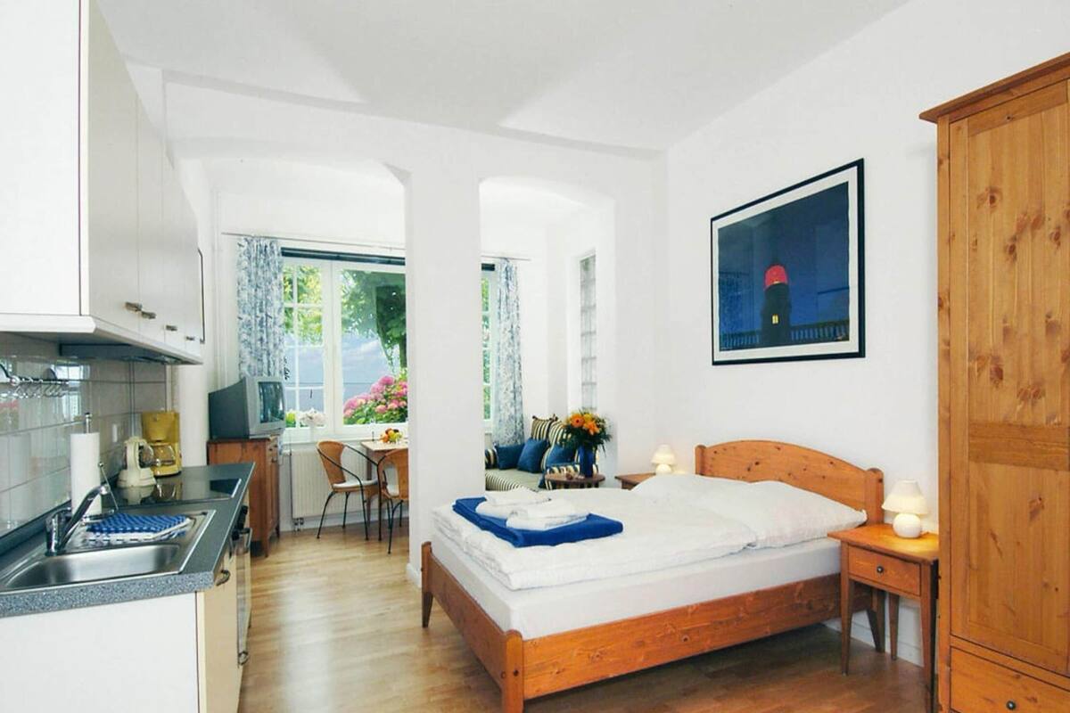 A bedroom with a wooden bed, a nightstand, a wooden wardrobe, a window with a view of flowers, and a kitchenette with a sink and stove.