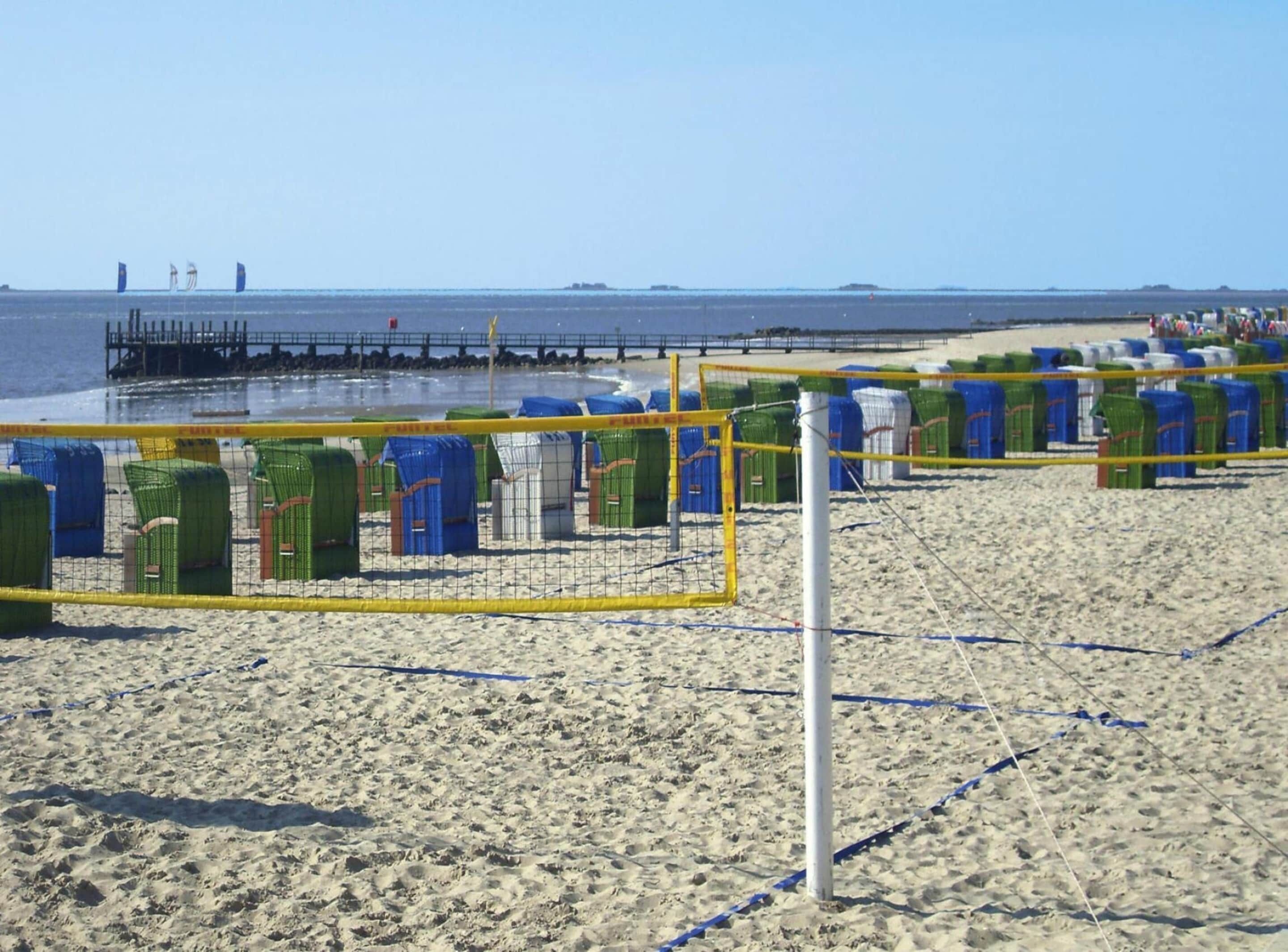 A beach volleyball court with net and chairs, a pier in the background, and the ocean.