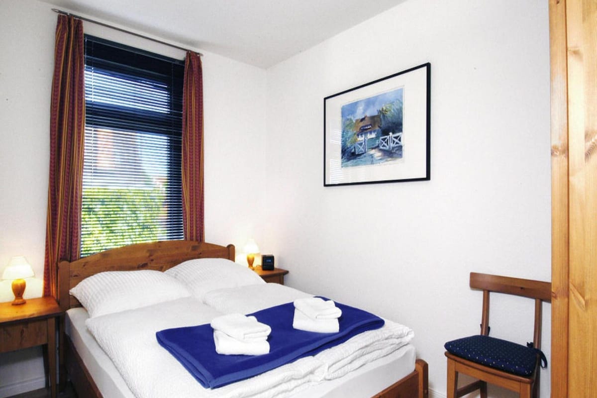 Ein ordentlich bezogenes Bett mit weißer und blauer Bettwäsche, ein gerahmtes Bild an der Wand und ein Holzstuhl.