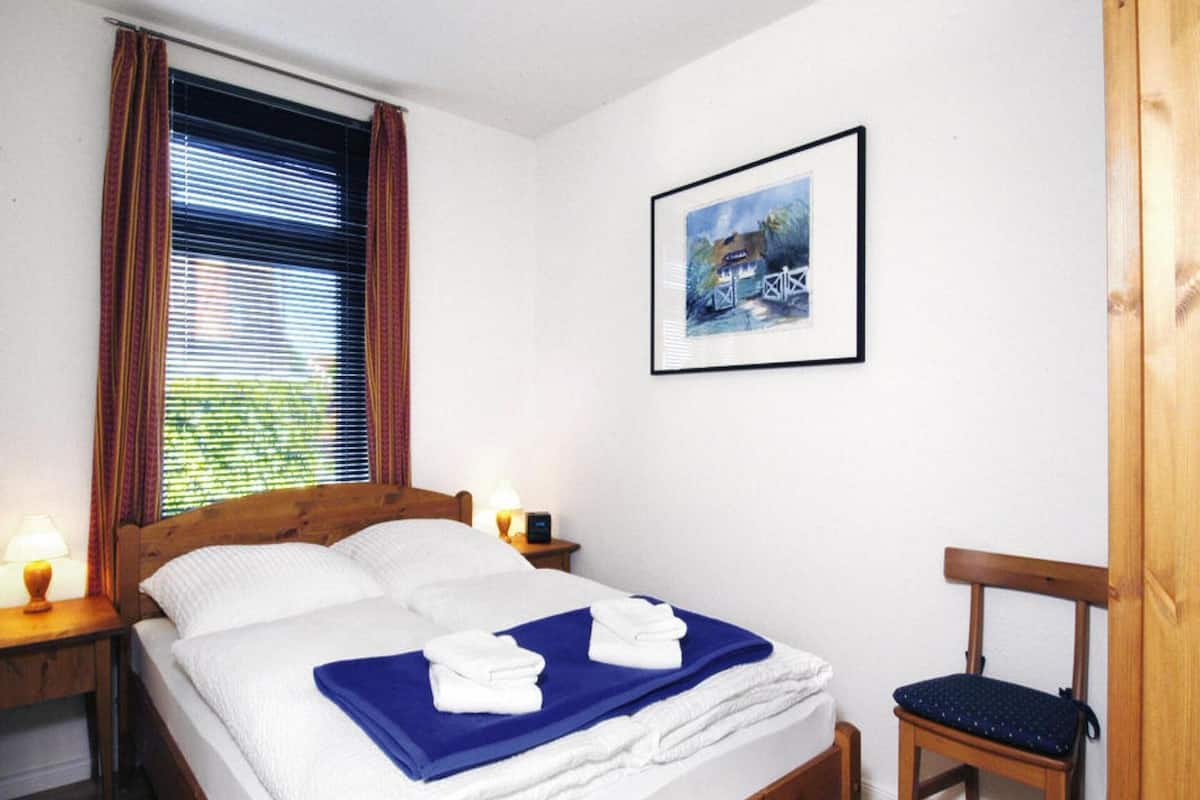 A neatly made bed with white and blue linens, a framed picture on the wall, and a wooden chair.