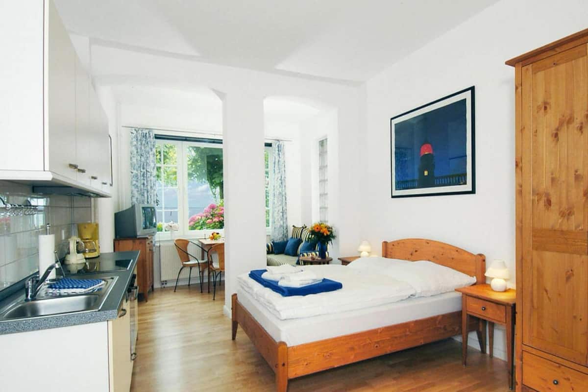 A bedroom with a wooden bed, a nightstand, a window with curtains, a dining area with a table and chairs, and a kitchen area with a sink and cabinets.