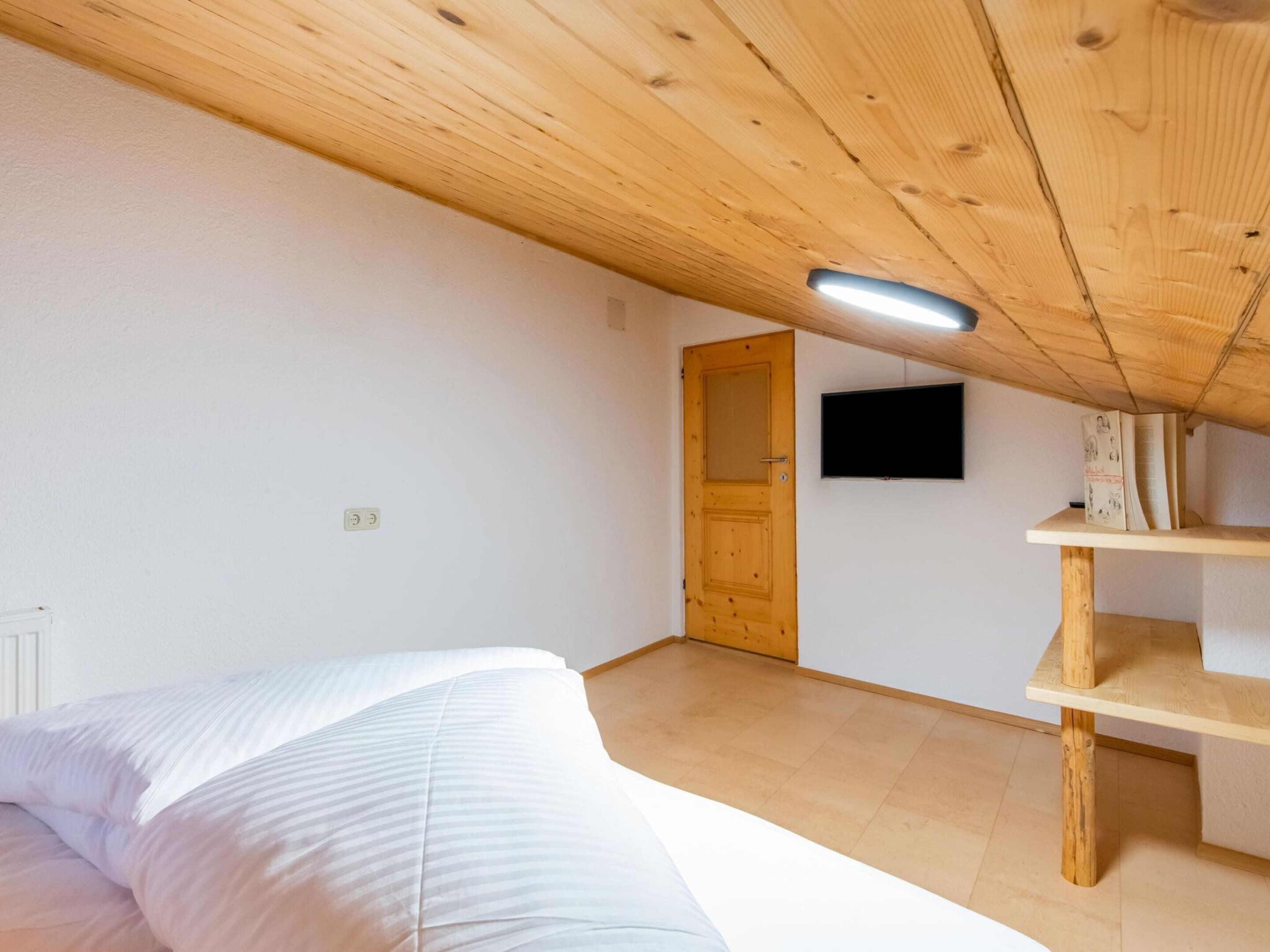 Ein Schlafzimmer mit Holzboden, einem Bett mit weißer Bettwäsche, einem Holzregal und einem Fernseher an der Wand.
