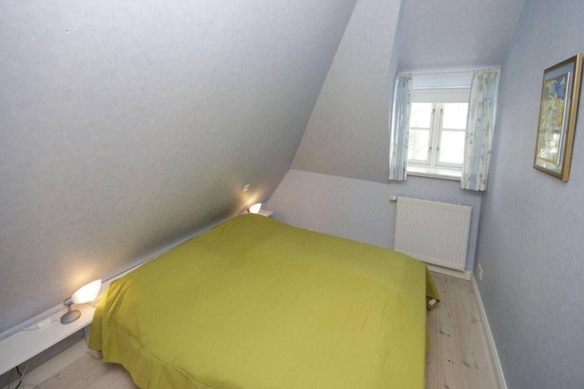 Ein Schlafzimmer mit schrägverdeckter Decke, einem Bett mit grüner Bettdecke, einer Nachttischlampe, einem Fenster mit Vorhängen und einer Heizung.