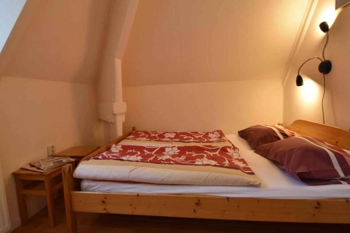 Una habitación pequeña y acogedora con techo inclinado, una cama individual con colcha estampada, una mesita de noche con lámpara y un aplique de pared.